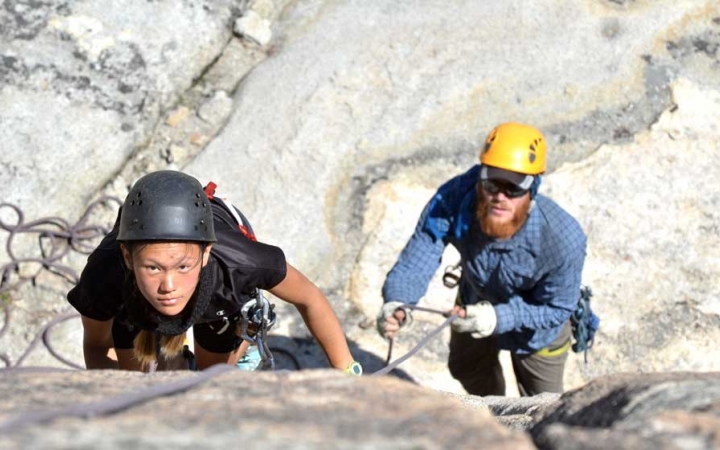 A student wearing safety gear looks up at the camera while they climb a rock wall. An instructor is below, belaying them.